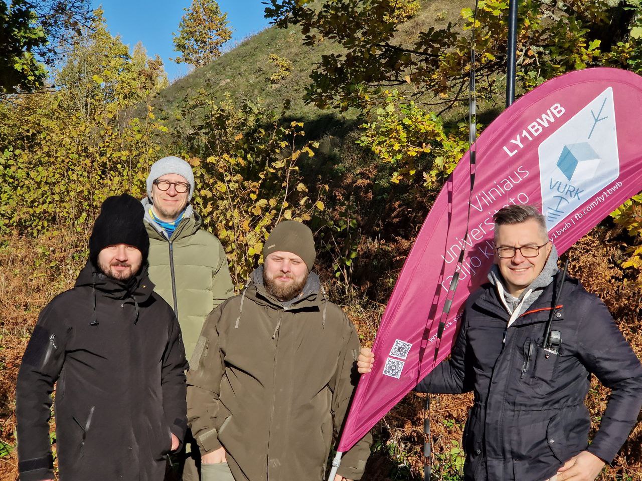 Keturi VU radijo klubo nariai stovi prie piliakalnio ir šypsosi. Dešinėje Simonas laiko violetinę klubo vėliavą. Fone spalvoti rudeniniai medžių lapai.