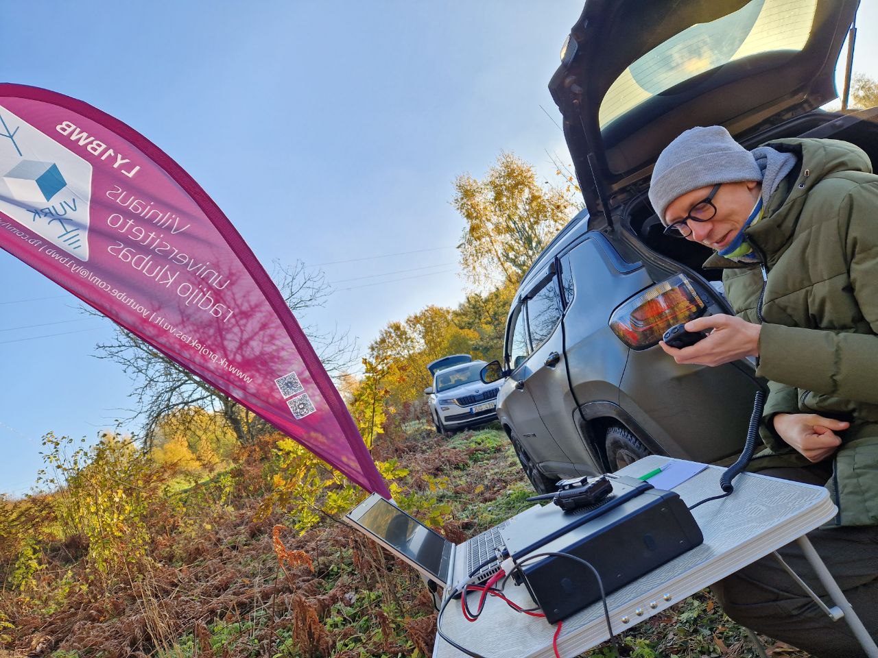 Juozas sėdi prie kelioninio staliuko ant kurio padėtas radijo aparatas ir kompiuteris. Rankoje laiko radijo aparato mikrofoną. Fone du automobiliai, rudeniškas peizažas, bei violetinė klubo vėliava